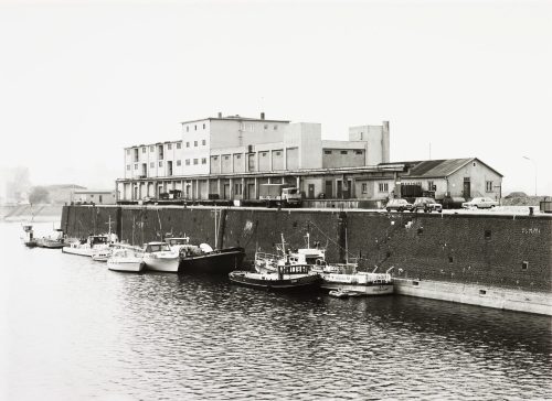 Tata Ronkholz (Fotograf*in), Binnenschiffe und Boote an einer Hafenbeckenmauer, vor dem Rhenania-Allgemeine-Gebäude im Hafen Düsseldorf, 1981