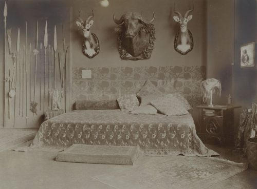 W. Hümmer (Künstler*in), Ohne Titel (Interieur der Wohnung eines Großwildjägers mit Wandtrophäen: Gazellen- und Büffelköpfe, an den Wänden drappierte Waffen und Wurfgeschosse), ca. 1910